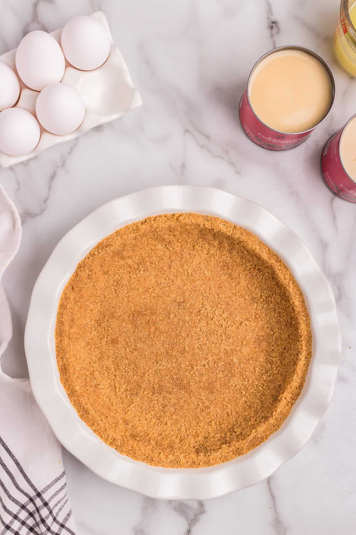 A graham cracker crust in a white ceramic pie dish sits on a marble countertop, ready for filling. Surrounding the crust are key ingredients for lemon pie, including a carton of eggs, open cans of sweetened condensed milk, and a measuring cup with additional ingredients. A striped kitchen towel is partially visible on the left side.