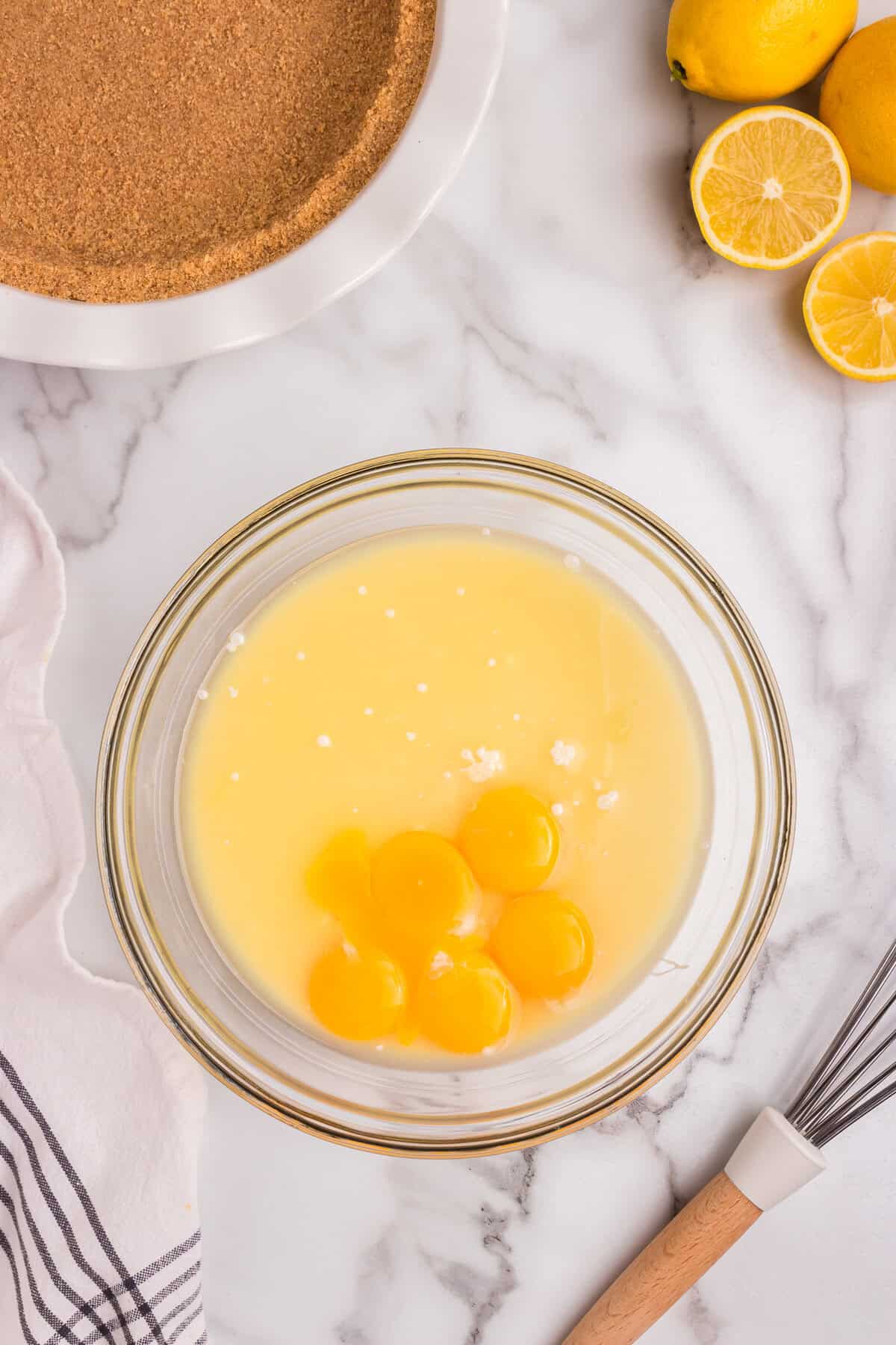 A glass mixing bowl filled with lemon pie ingredients, including egg yolks and sweetened condensed milk, sits on a marble countertop. A whisk with a wooden handle rests nearby, ready for mixing. In the background, a graham cracker crust in a white ceramic pie dish is prepared for filling. Fresh lemons, some whole and some sliced, are placed nearby. A striped kitchen towel is draped on the counter.