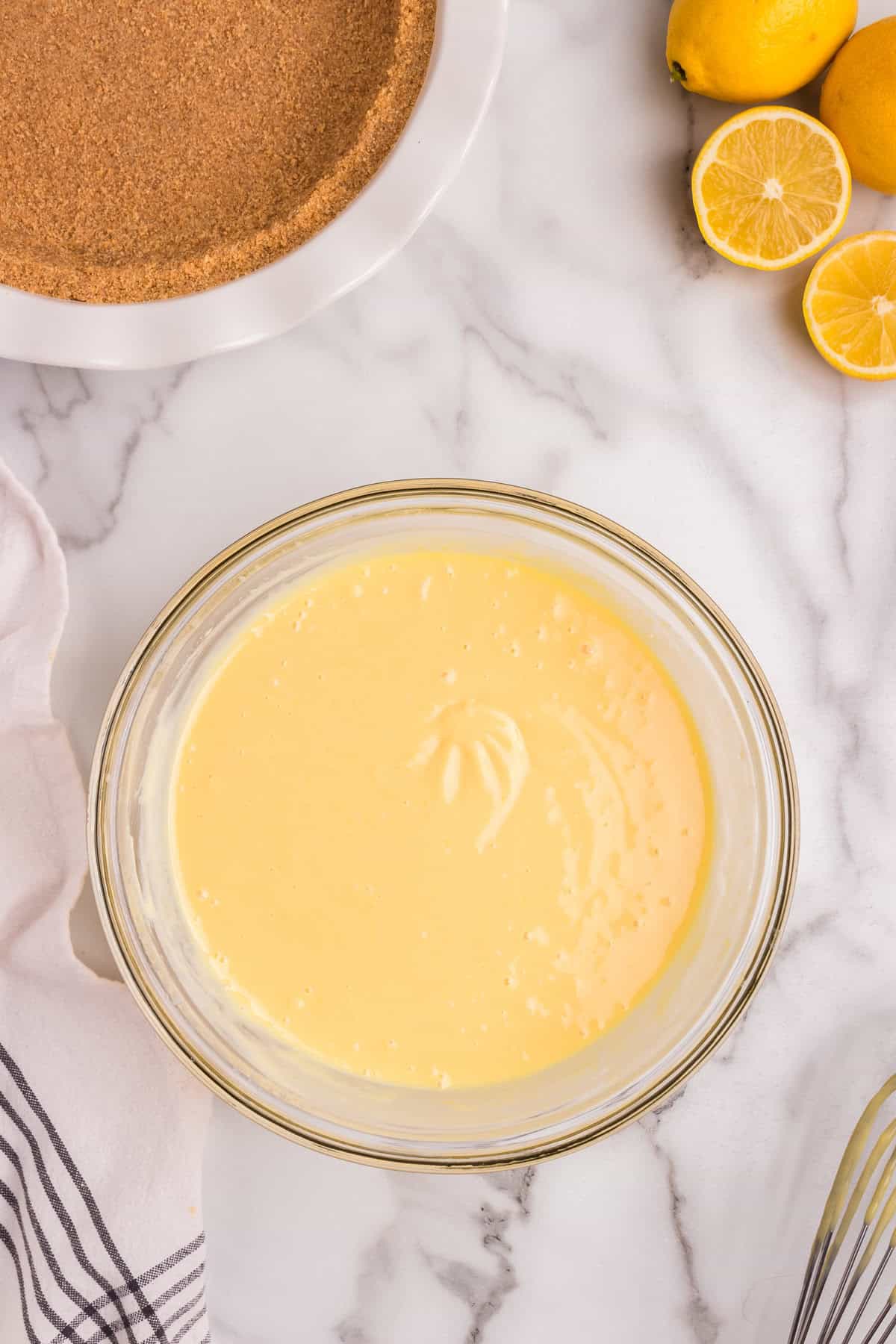 A glass mixing bowl filled with smooth, pale yellow lemon pie filling sits on a marble countertop. A whisk has left soft trails in the creamy mixture. In the background, a pre-baked graham cracker crust in a white ceramic pie dish is ready for filling. Fresh lemons, some whole and some sliced, rest nearby. A striped kitchen towel is draped on the counter.