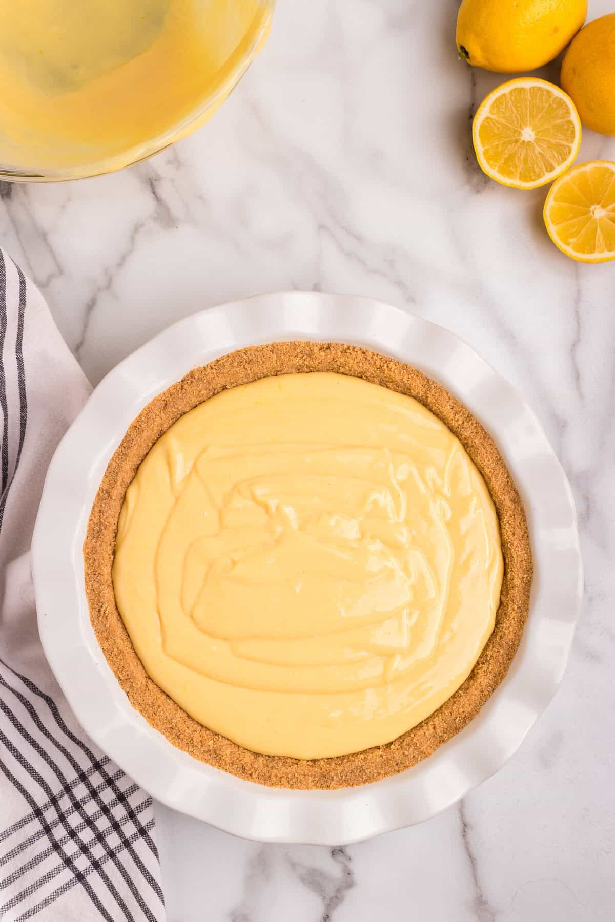 A freshly filled lemon pie with a slightly swirled, creamy yellow filling sits in a white ceramic pie dish. The graham cracker crust is evenly formed along the edges. A glass mixing bowl with remnants of the lemon filling is visible in the background, along with fresh lemons, some whole and some sliced. A striped kitchen towel rests on the marble countertop nearby.