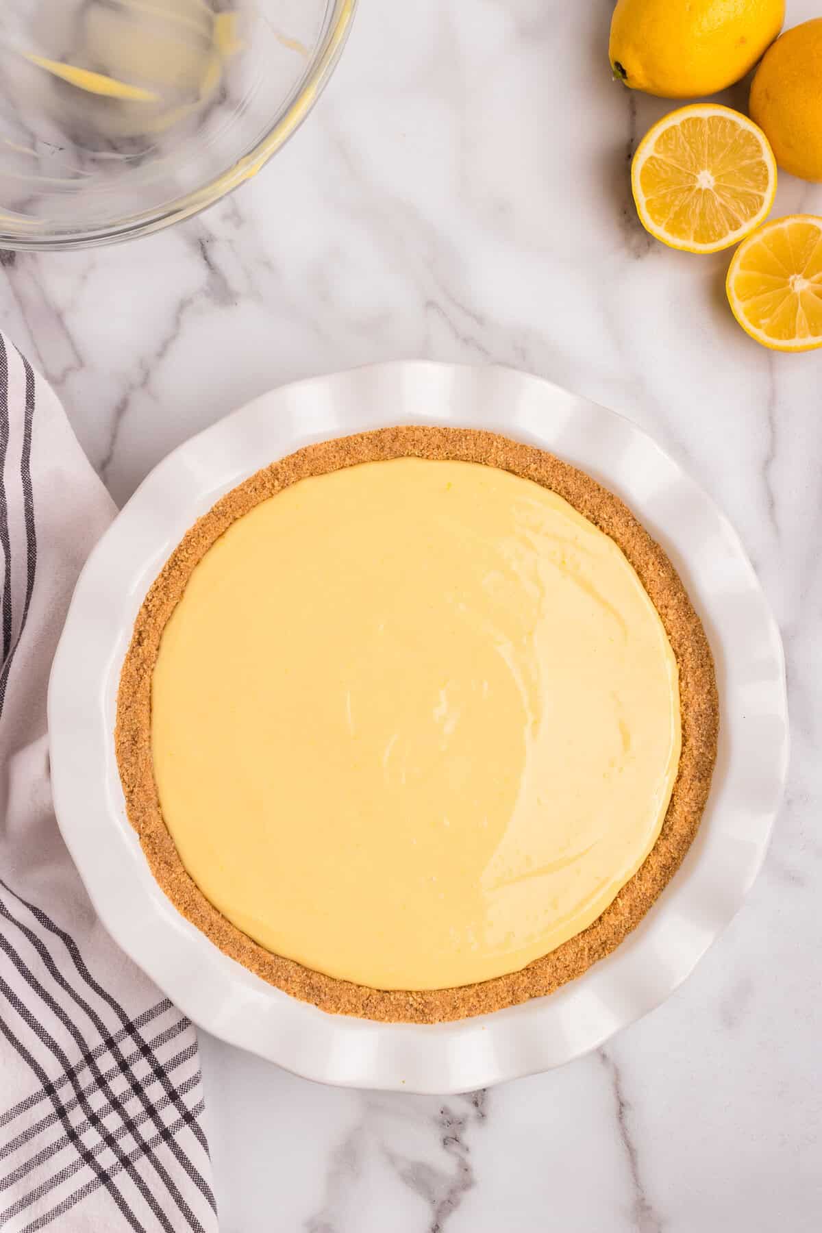 An unbaked lemon pie with a smooth, creamy yellow filling sits in a white ceramic pie dish. The graham cracker crust is evenly pressed along the edges. A glass mixing bowl with remnants of the filling is visible in the background, along with fresh lemons, some whole and some sliced. A striped kitchen towel is draped nearby on the marble countertop.