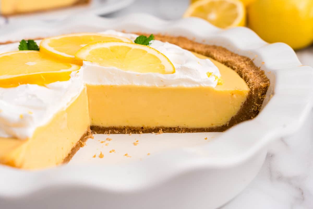 A close-up of a partially sliced lemon pie in a white ceramic dish, showing its smooth, creamy lemon filling and a golden graham cracker crust. The pie is topped with fluffy whipped cream, thin slices of fresh lemon, and a few mint leaves. Crumbs are scattered around the base, and fresh lemons are visible in the blurred background.