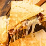 A close-up of a cheesy pulled pork quesadilla being lifted with a spatula. The golden-brown, crispy tortilla is filled with melted cheese and tender pulled pork, with gooey cheese stretching between the slices. The quesadilla sits on a wooden surface with additional pieces in the background.
