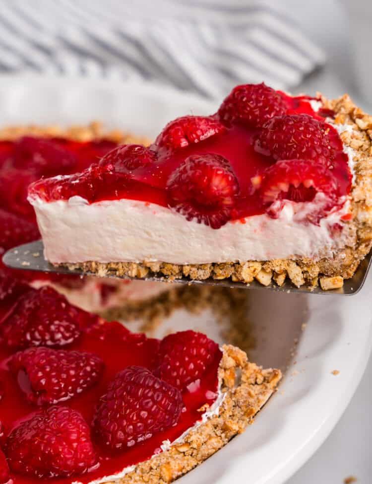 A close-up of a slice of raspberry pretzel pie being lifted from a white pie dish. The dessert features a crunchy pretzel crust, a creamy white filling, and a glossy raspberry topping with whole raspberries. A striped kitchen towel is visible in the blurred background.