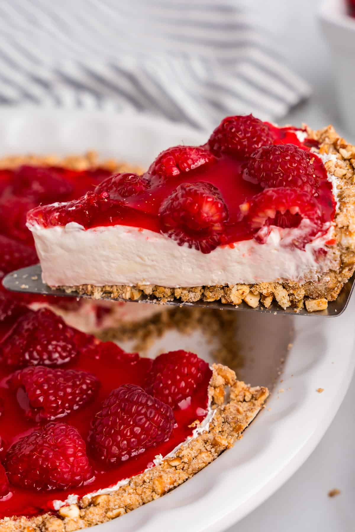 A close-up of a slice of raspberry pretzel pie being lifted from a white pie dish. The dessert features a crunchy pretzel crust, a creamy white filling, and a glossy raspberry topping with whole raspberries. A striped kitchen towel is visible in the blurred background.