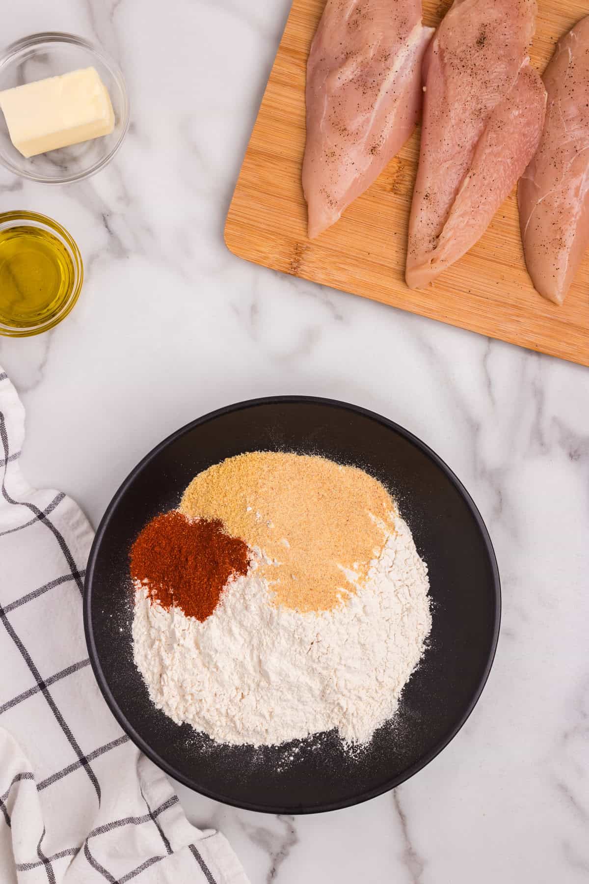 Overhead picture of smothered chicken flour mixture on a black plate, wooden cutting board with raw chicken and oil.