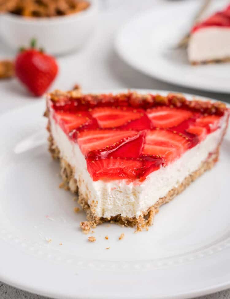 A close-up of a slice of strawberry pretzel pie on a white plate. The slice has a crunchy pretzel crust, a thick creamy white filling, and a glossy red strawberry topping with fresh sliced strawberries. In the background, a whole pie, a fresh strawberry, and a bowl of pretzels are slightly blurred.