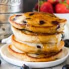 Stack of blueberry pancakes with syrup being poured over them on white plate