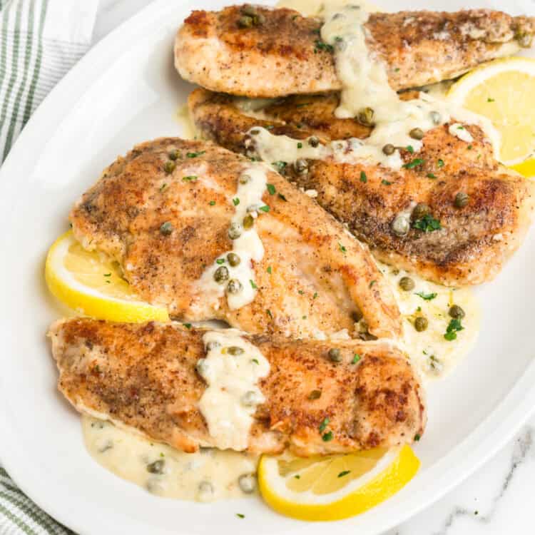 A white serving platter with golden-brown chicken piccata fillets drizzled with a creamy lemon and caper sauce. The dish is garnished with fresh parsley and lemon slices, creating a vibrant and appetizing presentation.