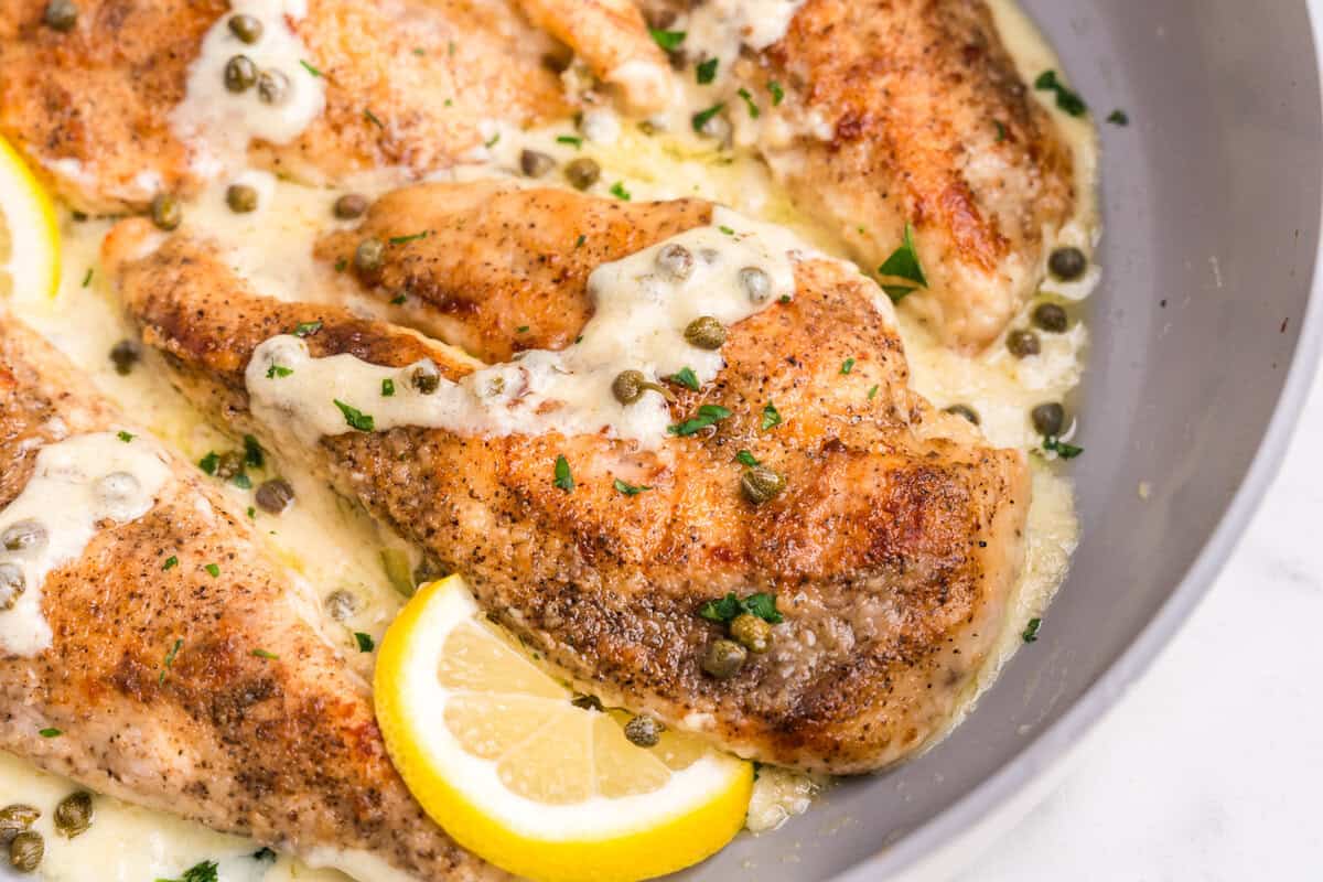 A close-up of chicken piccata fillets in a creamy lemon caper sauce, garnished with fresh parsley. The golden-brown chicken is nestled in the sauce, with slices of bright yellow lemon adding a fresh contrast. The capers are sprinkled over the dish, enhancing its vibrant and tangy presentation.





