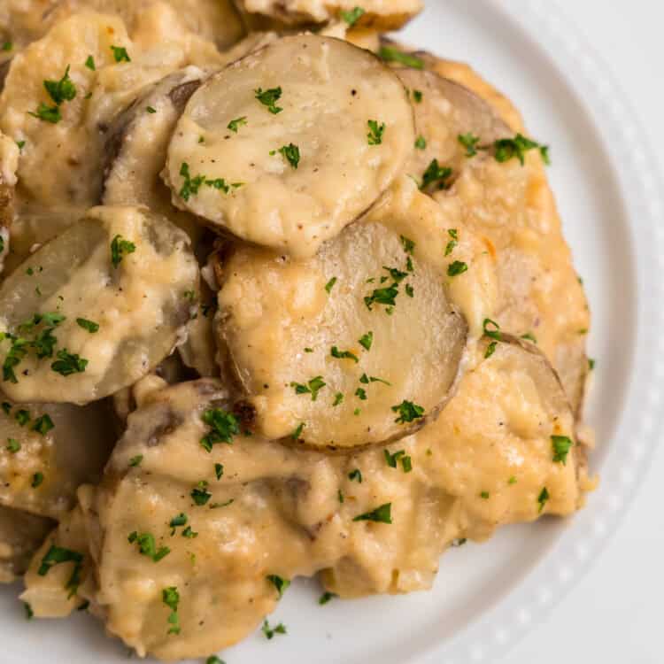 A close-up of a serving of creamy au gratin potatoes on a white plate, garnished with freshly chopped parsley. The dish features tender, thinly sliced potatoes coated in a rich, cheesy sauce with a golden hue. The vibrant green parsley adds a fresh contrast to the warm, comforting meal.