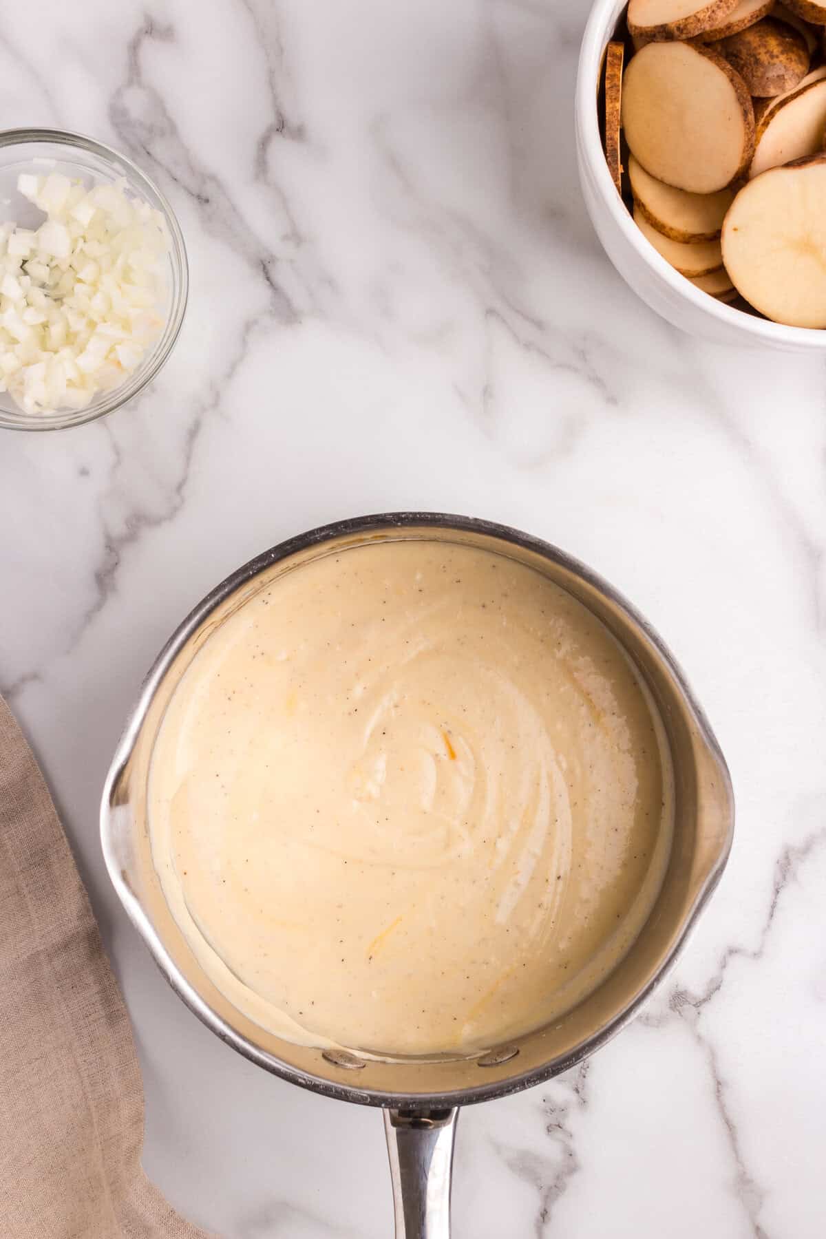 A stainless steel saucepan filled with a smooth, creamy cheese sauce with specks of seasoning. The sauce has a rich, velvety texture, likely intended for a potato-based dish. Surrounding the saucepan are a bowl of sliced potatoes and a bowl of diced onions, all arranged on a white marble countertop, indicating ongoing meal preparation.
