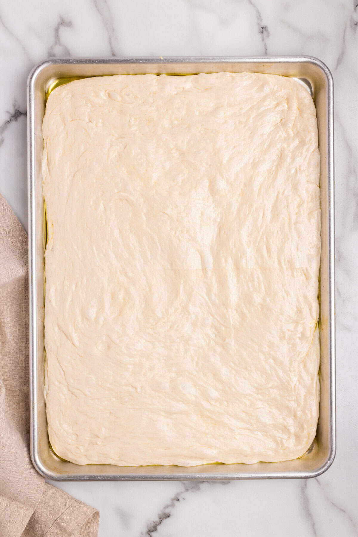 The dough has been left to proof in the baking sheet and has expanded, filling the pan more fully. A light layer of olive oil pools around the edges, giving the dough a glossy appearance.