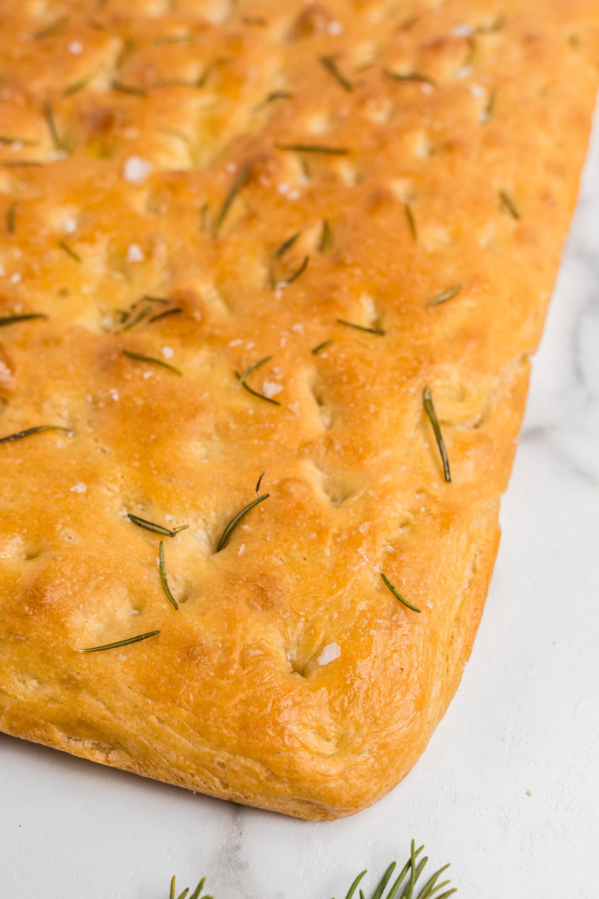 A close-up of the baked focaccia, highlighting the crisp, golden crust. The fresh rosemary is slightly toasted, and flakes of coarse salt add texture and contrast.