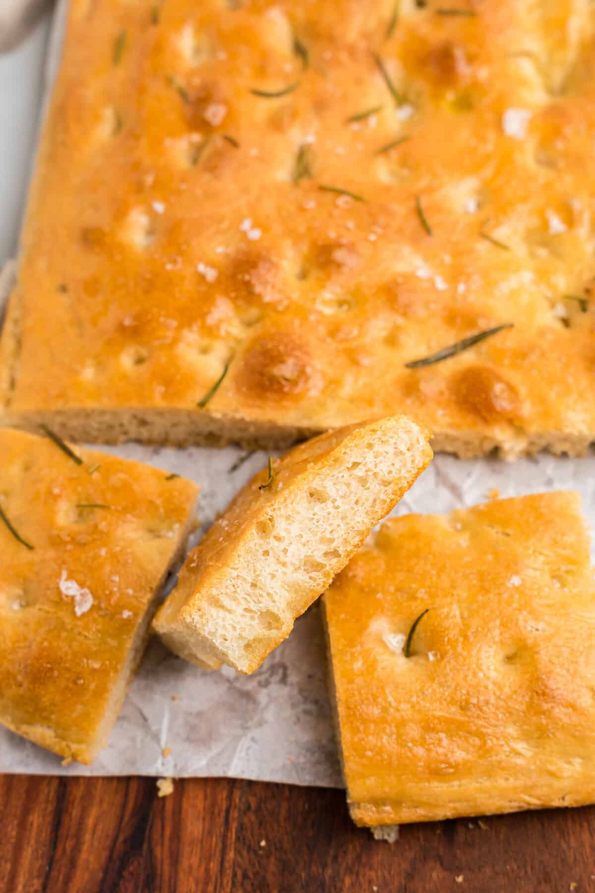 The focaccia has been cut into square pieces and placed on a sheet of parchment paper atop a wooden cutting board. One piece is slightly lifted to show its fluffy, airy interior.