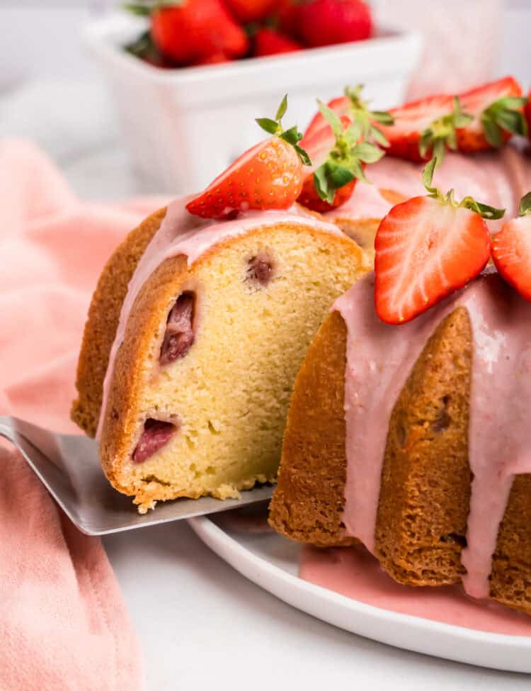 A slice of strawberry pound cake being lifted from the whole cake with a cake server. The cake has a golden brown crust, a soft interior with baked-in strawberry pieces, and is topped with pink strawberry glaze and fresh strawberry slices. In the background, a container of fresh strawberries and a pink cloth napkin are placed on a marble countertop.