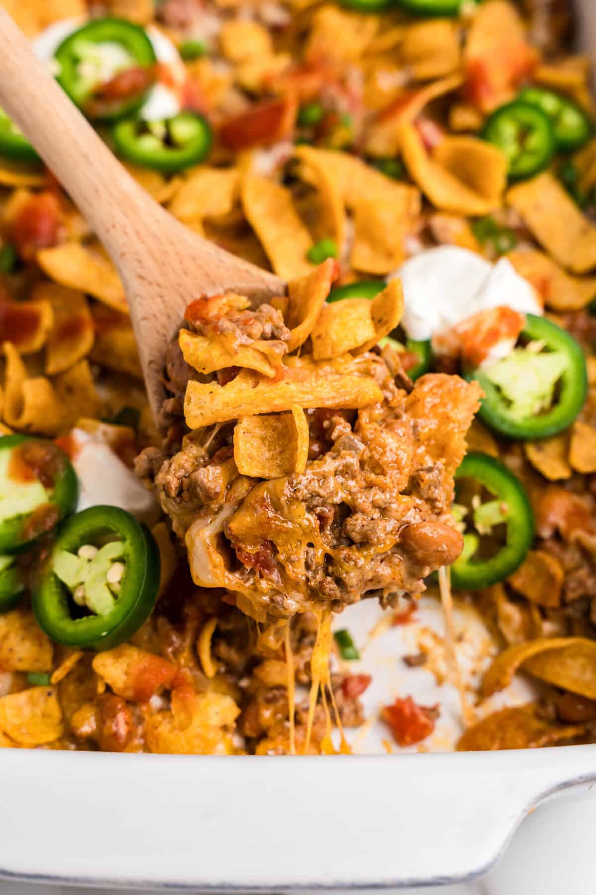 A close-up of a wooden spoon scooping a portion of walking taco casserole from a white baking dish. The casserole consists of seasoned ground beef, melted cheese, Fritos corn chips, diced tomatoes, pinto beans, and is garnished with sliced jalapeños and dollops of sour cream. The melted cheese stretches as the portion is lifted, showing the layers of ingredients.