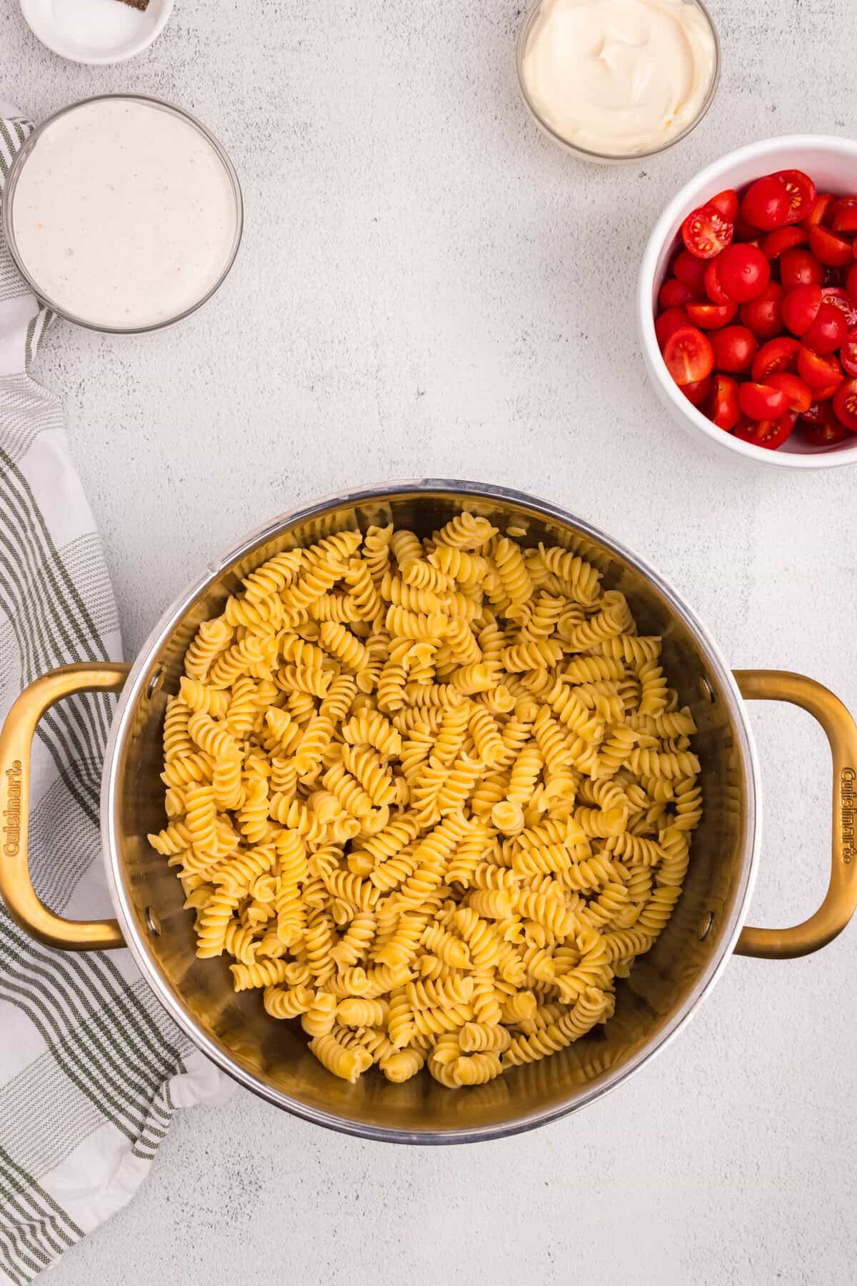 verhead view of a pot filled with cooked rotini pasta, surrounded by bowls containing halved cherry tomatoes, mayonnaise, creamy dressing, and seasonings on a white countertop.