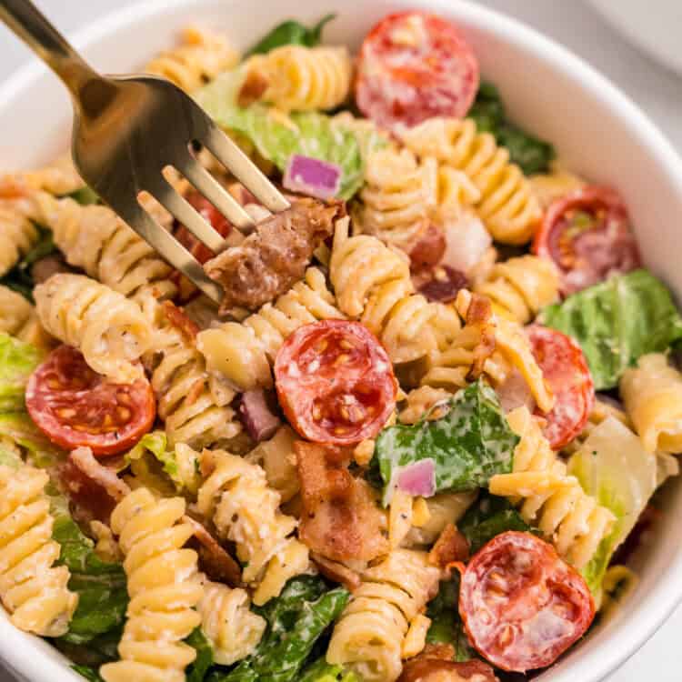 A close-up of a bowl of BLT pasta salad, with a gold fork spearing a piece of bacon. The salad features rotini pasta, halved cherry tomatoes, romaine lettuce, red onion, shredded cheddar, and creamy dressing.
