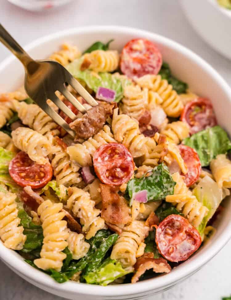 A close-up of a bowl of BLT pasta salad, with a gold fork spearing a piece of bacon. The salad features rotini pasta, halved cherry tomatoes, romaine lettuce, red onion, shredded cheddar, and creamy dressing.