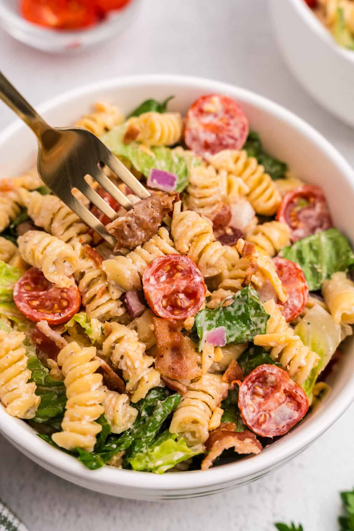 A close-up of a bowl of BLT pasta salad, with a gold fork spearing a piece of bacon. The salad features rotini pasta, halved cherry tomatoes, romaine lettuce, red onion, shredded cheddar, and creamy dressing.