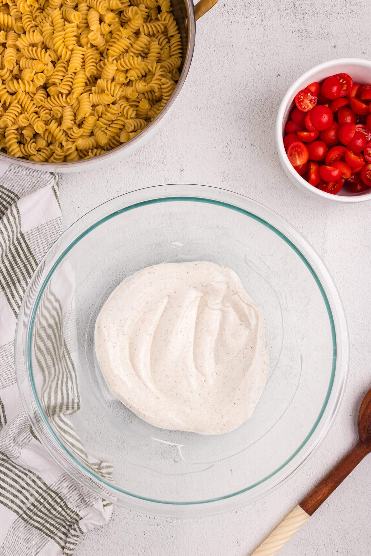 Overhead view of a glass mixing bowl with creamy seasoned dressing, next to a pot of cooked rotini pasta and a bowl of halved cherry tomatoes on a white countertop.