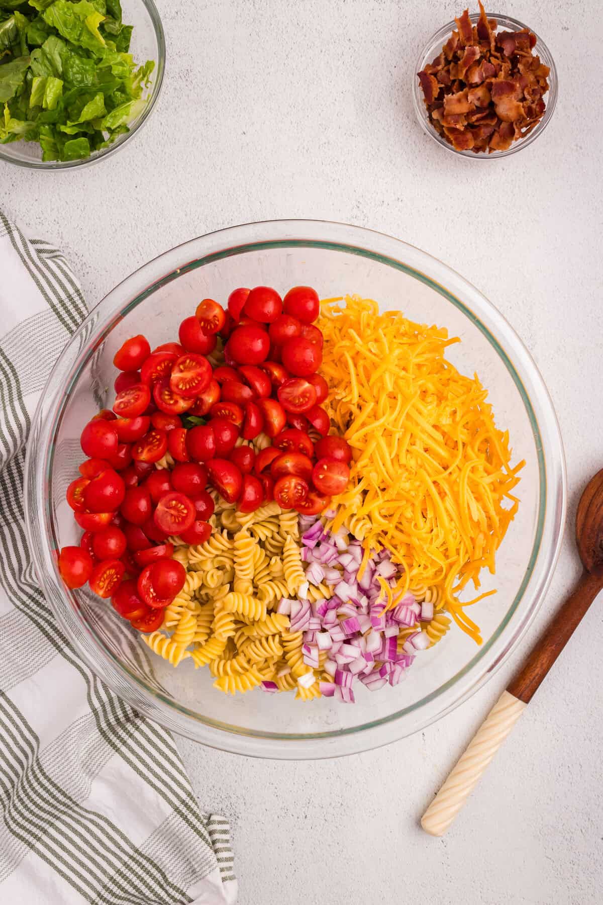 Overhead view of a glass mixing bowl filled with uncooked rotini pasta, halved cherry tomatoes, shredded cheddar cheese, and diced red onion, with bowls of chopped lettuce and crumbled bacon nearby on a white surface.