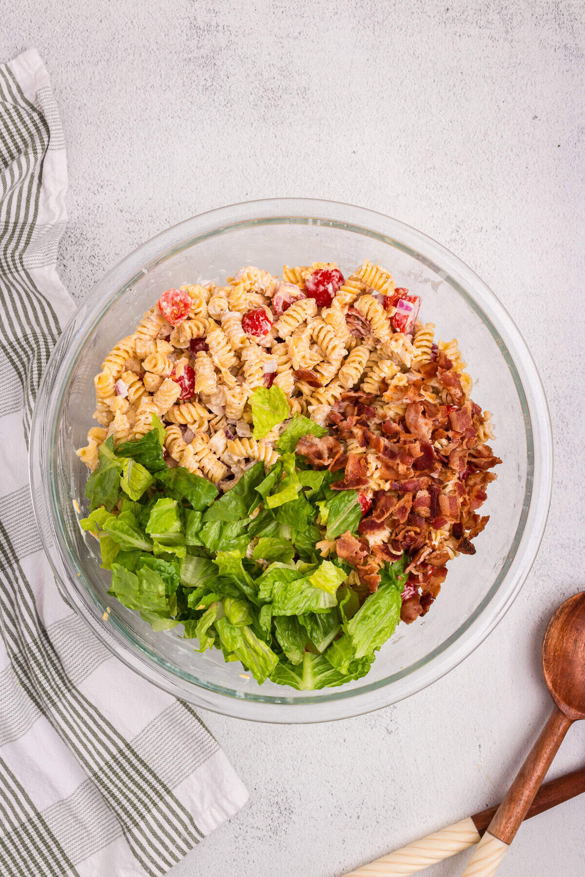 Overhead view of a glass bowl filled with creamy rotini pasta salad, topped with chopped romaine lettuce and crispy crumbled bacon, ready to be mixed together.