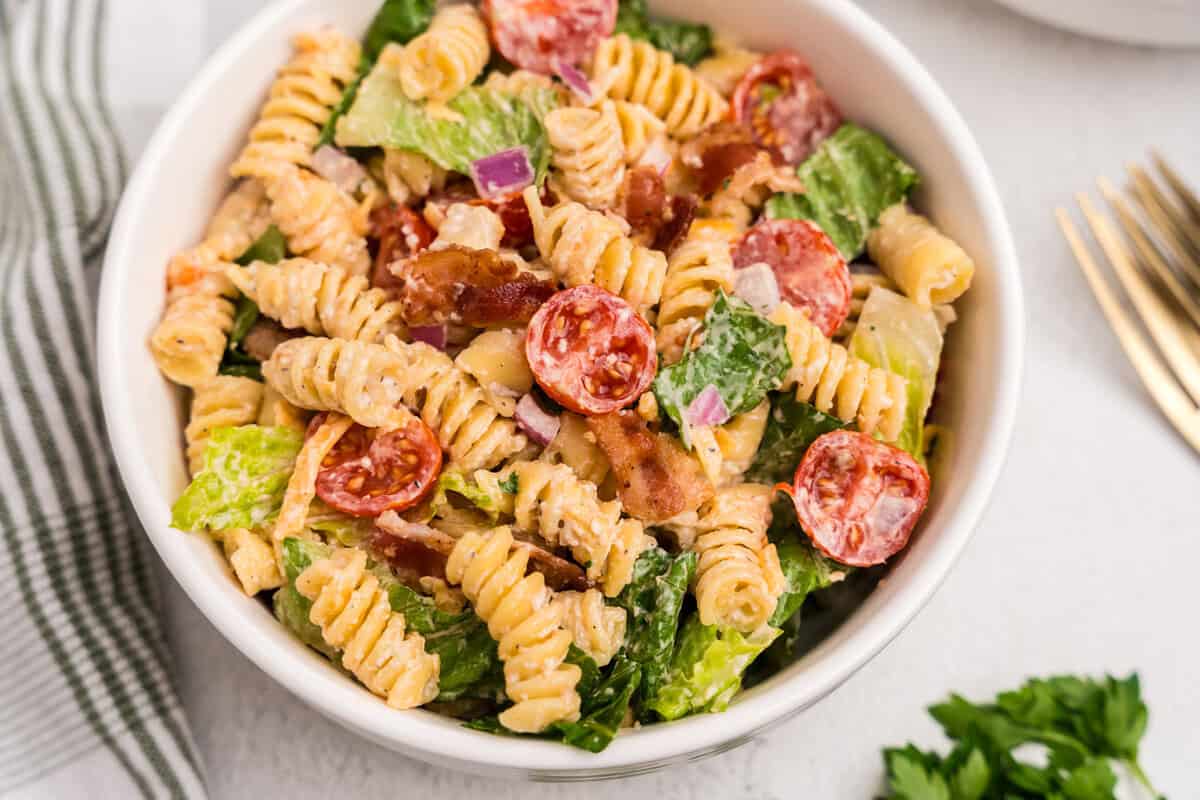A serving of the BLT pasta salad in a white bowl, beautifully presented with colorful ingredients and garnished with chopped parsley. Gold forks and parsley leaves lie nearby for garnish and serving.