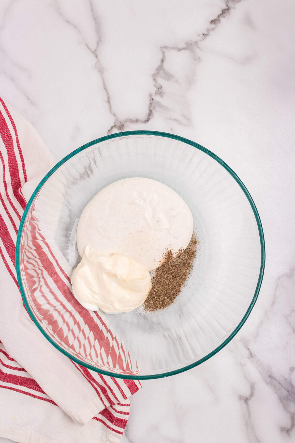 A glass mixing bowl on a marble surface with mayonnaise, sour cream, and freshly ground black pepper, ready to be mixed. A red-and-white striped towel lies beside the bowl.
