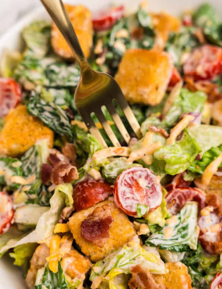 A zoomed-in view of a serving fork diving into the vibrant salad, emphasizing the textures of the croutons, crisp lettuce, tomatoes, bacon, and dressing.