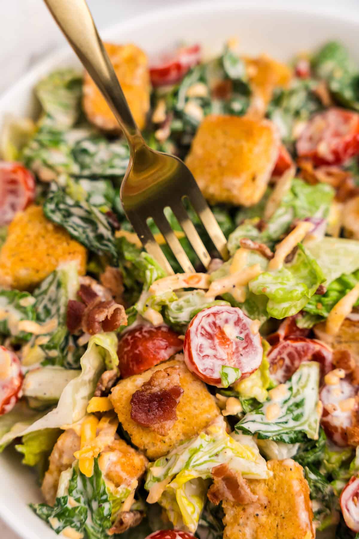 A zoomed-in view of a serving fork diving into the vibrant salad, emphasizing the textures of the croutons, crisp lettuce, tomatoes, bacon, and dressing.