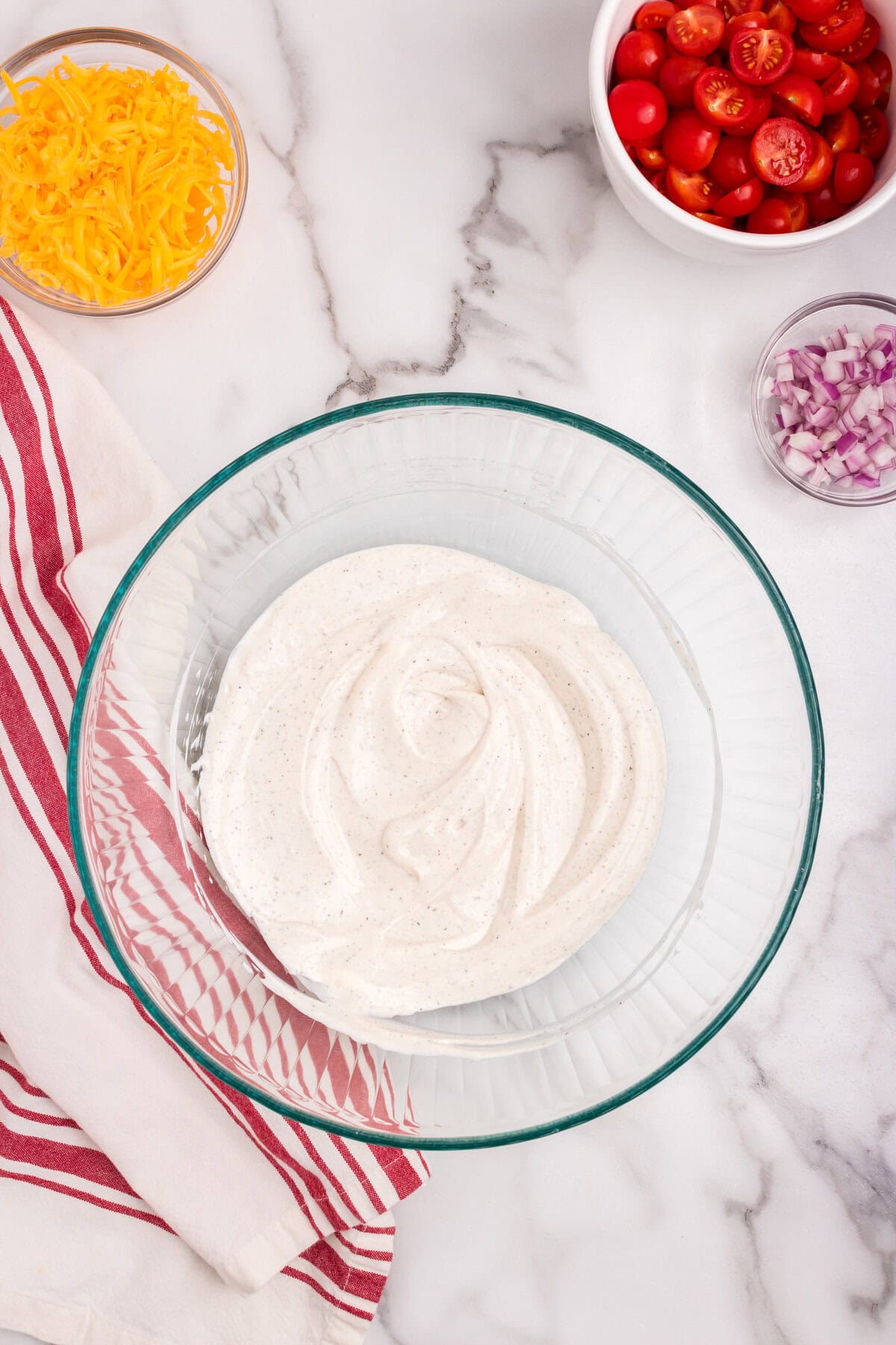 The bowl now contains a smooth, creamy dressing mixture. In the background, shredded cheddar cheese, halved cherry tomatoes, and chopped red onions are prepped in small bowls.