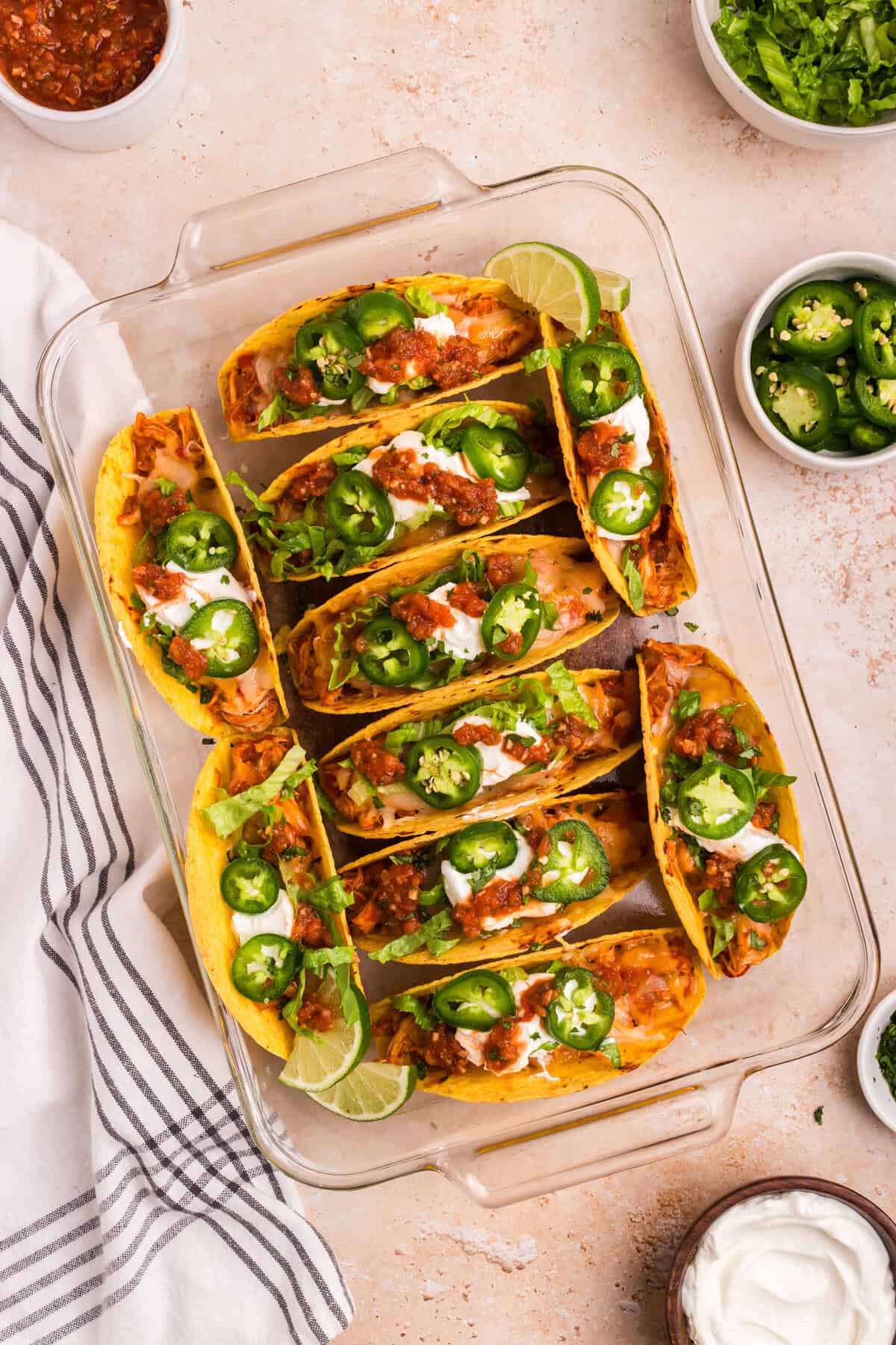 A glass baking dish filled with fully dressed baked chicken tacos, topped with sour cream, salsa, sliced jalapeños, and shredded lettuce. Lime wedges, fresh cilantro, and bowls of toppings surround the dish.