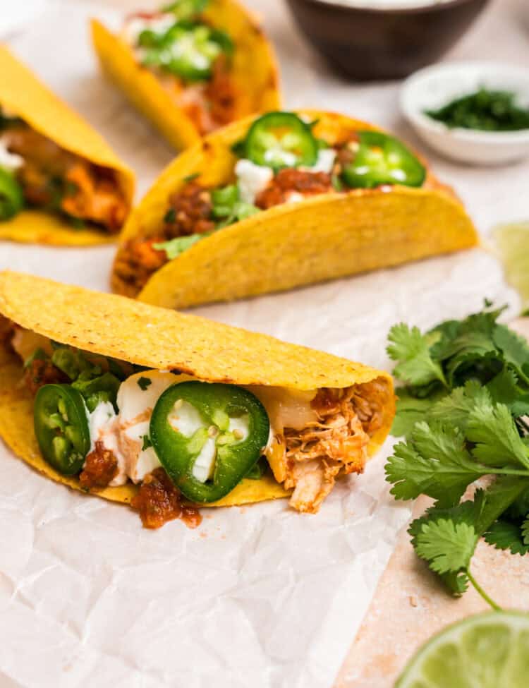 A few fully assembled baked chicken tacos resting on parchment paper, garnished with jalapeño slices, sour cream, salsa, and lettuce. Fresh cilantro and lime slices add a pop of color nearby.