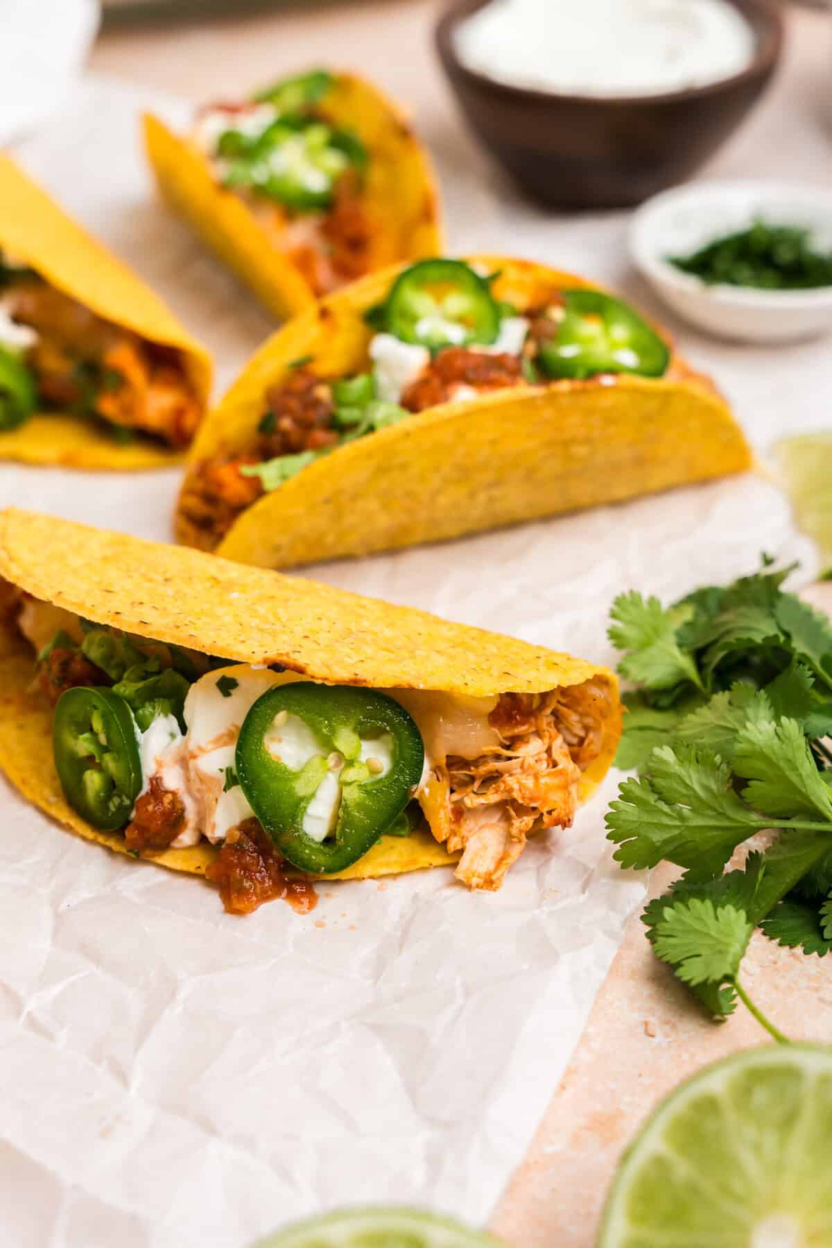 A few fully assembled baked chicken tacos resting on parchment paper, garnished with jalapeño slices, sour cream, salsa, and lettuce. Fresh cilantro and lime slices add a pop of color nearby.