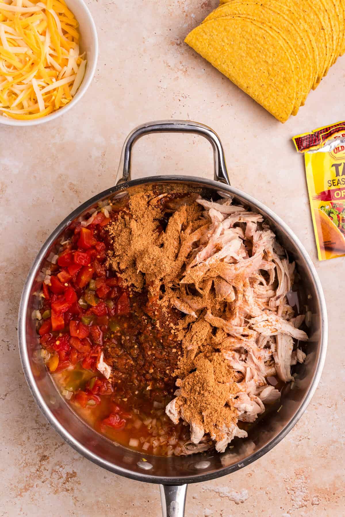 The same skillet from the first image, now with taco seasoning sprinkled over the shredded chicken mixture, preparing to be stirred together. A packet of taco seasoning is seen nearby.