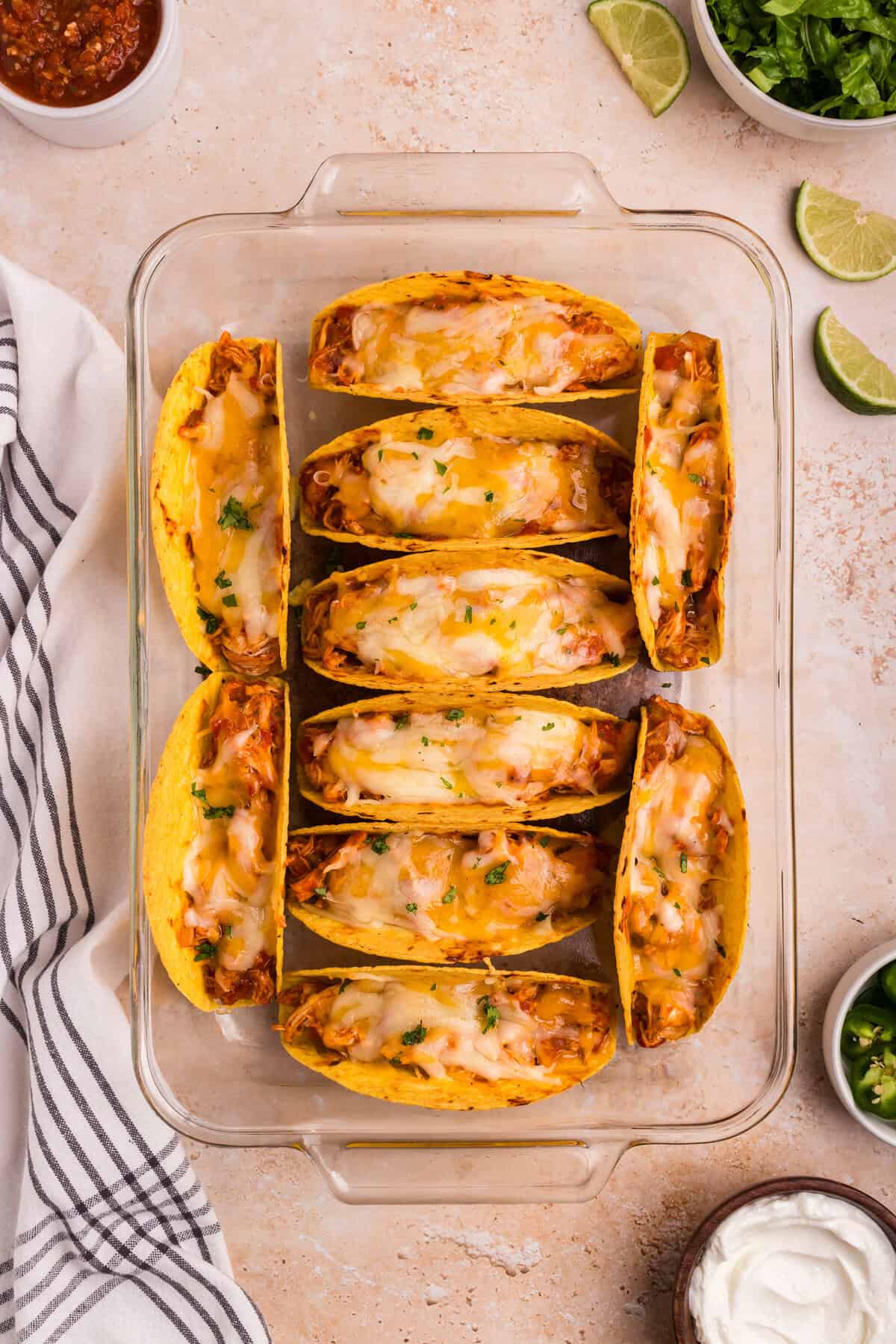 The tacos are baked, the cheese is melted and slightly golden, and the dish is garnished with fresh chopped herbs. Surrounding elements include lime wedges, sour cream, and jalapeño slices