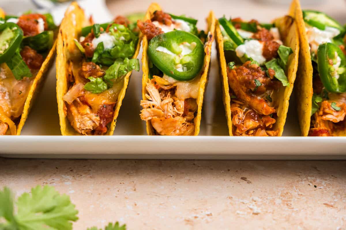 A row of five baked chicken tacos served on a white platter, filled with shredded chicken, melted cheese, lettuce, sour cream, salsa, and sliced jalapeños. The tacos are neatly arranged and vibrant with toppings.