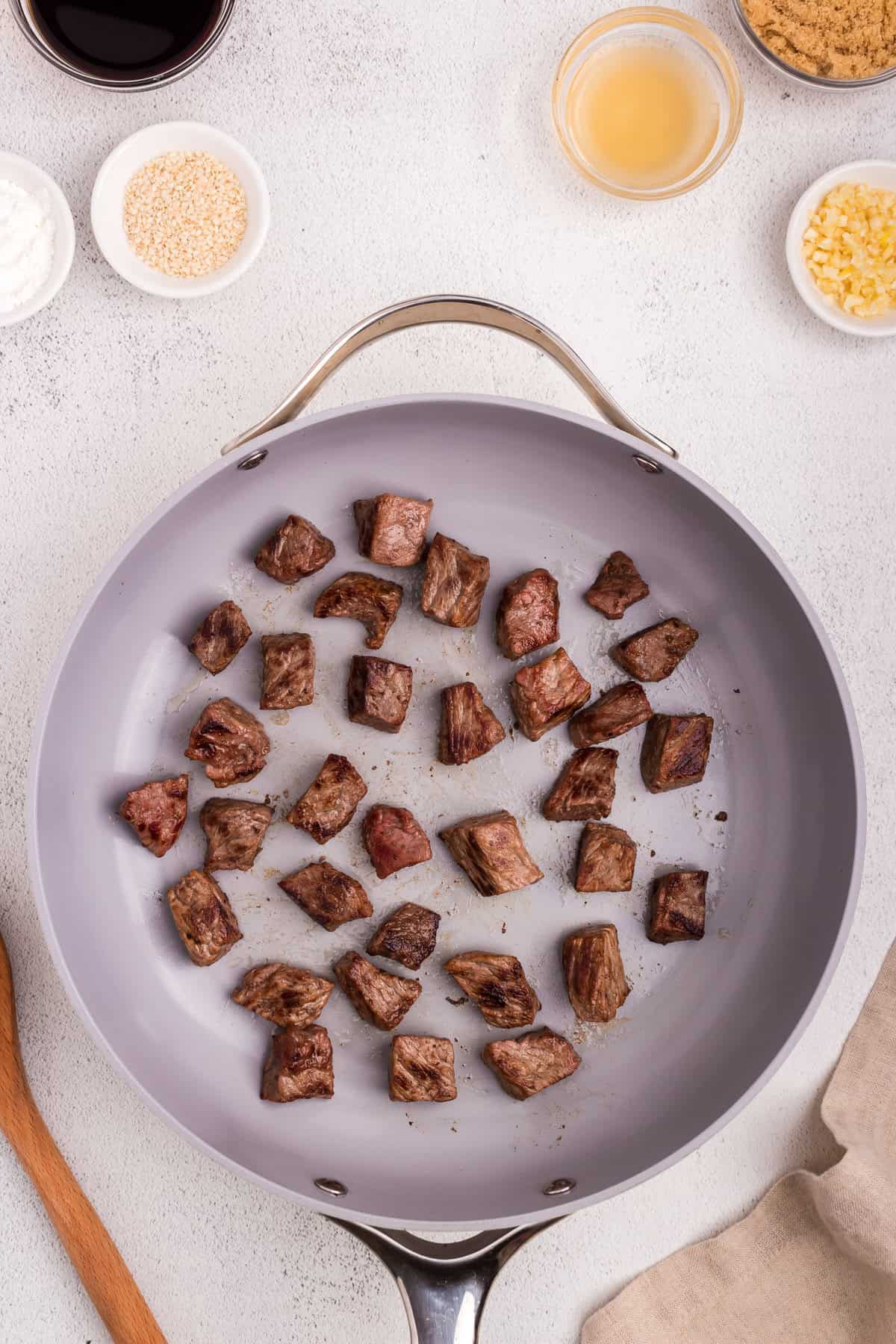 Cubes of beef searing in a light-colored skillet; surrounding ingredients like soy sauce, sesame seeds, minced garlic, brown sugar, and cornstarch are arranged on the counter.