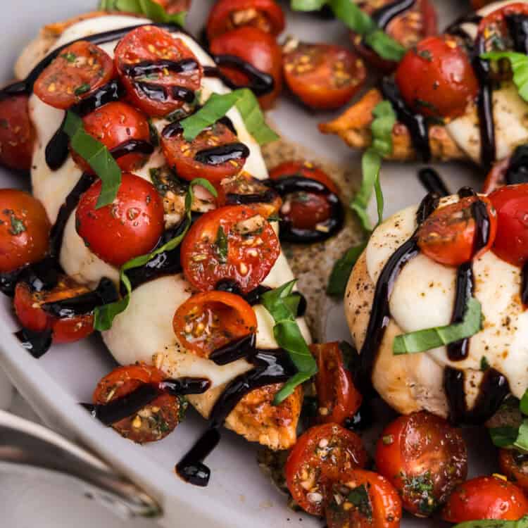Close-up of bruschetta chicken breasts topped with melted mozzarella, halved cherry tomatoes, fresh basil ribbons, and drizzled with balsamic glaze in a white skillet.