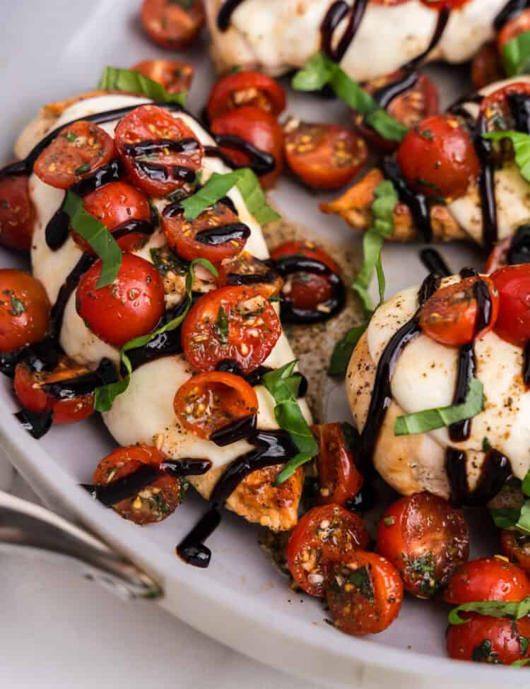 Close-up of bruschetta chicken breasts topped with melted mozzarella, halved cherry tomatoes, fresh basil ribbons, and drizzled with balsamic glaze in a white skillet.
