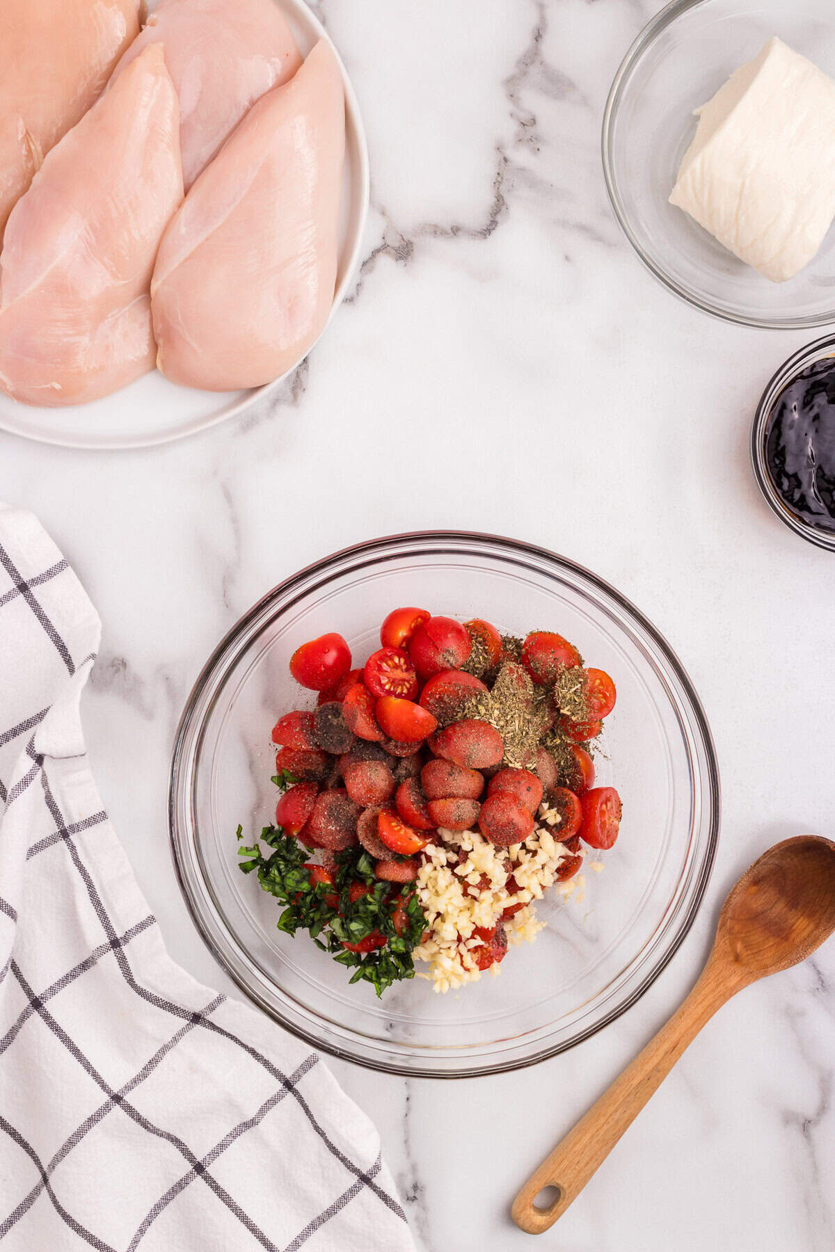 A bowl with halved cherry tomatoes, minced garlic, chopped basil, and Italian seasoning sits beside a plate of raw chicken breasts, a ball of mozzarella, and a small bowl of balsamic glaze—ingredients for Bruschetta Chicken.