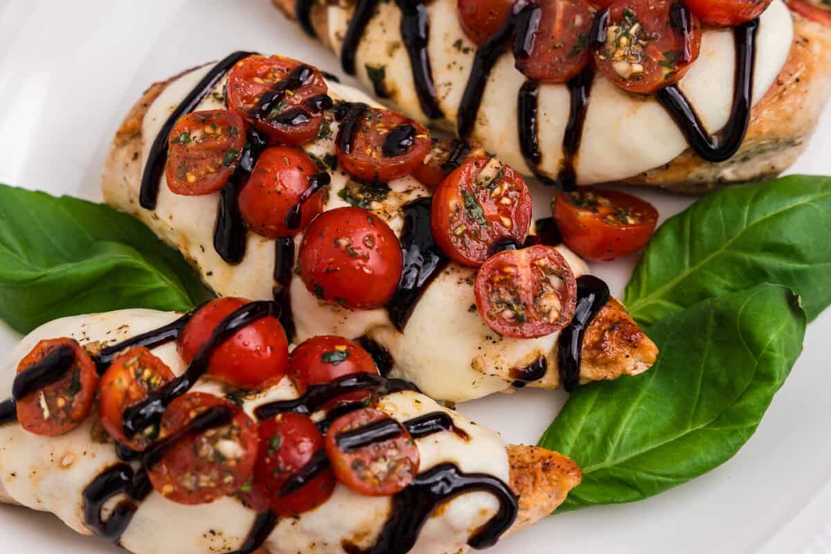 Overhead close-up of three pieces of Bruschetta Chicken on a white plate with basil leaves, showing the colorful contrast of fresh tomatoes, dark balsamic glaze, and creamy mozzarella.