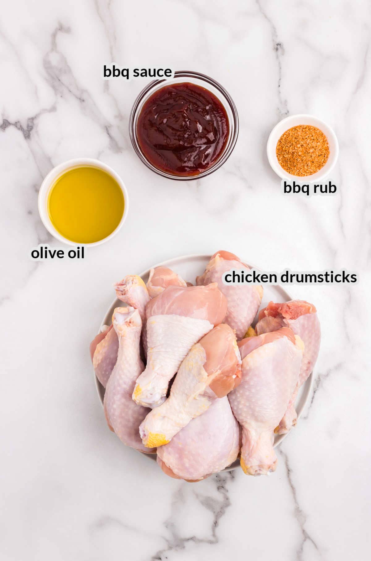 Raw chicken drumsticks, olive oil, BBQ sauce, and BBQ rub on a marble countertop.