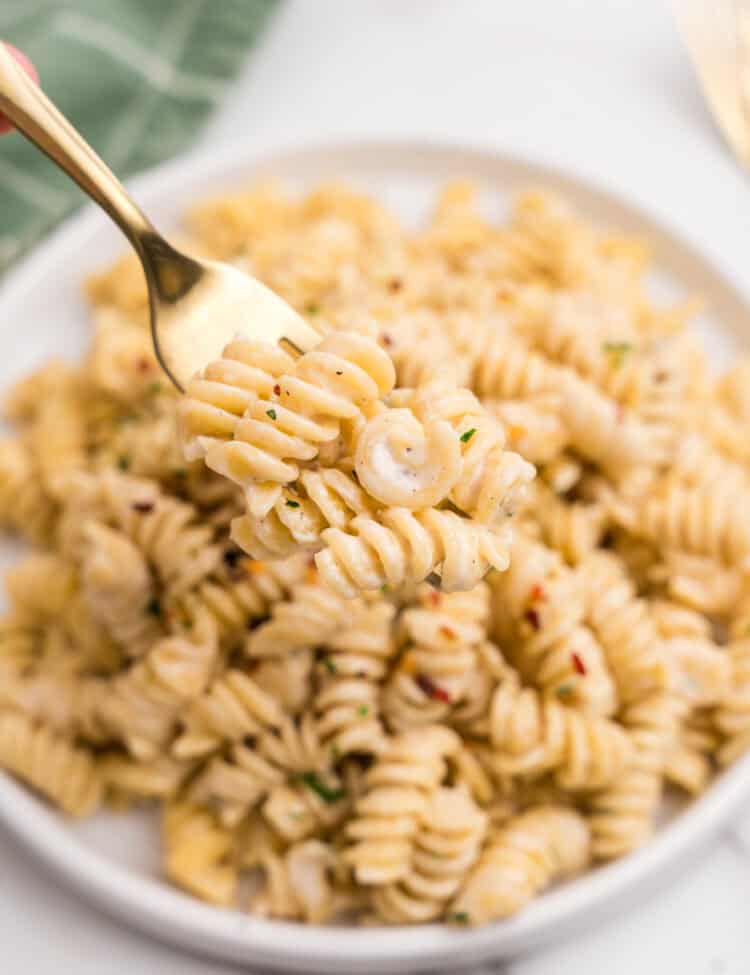A fork lifting a bite of creamy rotini pasta from a plate, showing the cheesy sauce coating the noodles, with red pepper flakes and parsley as garnish.