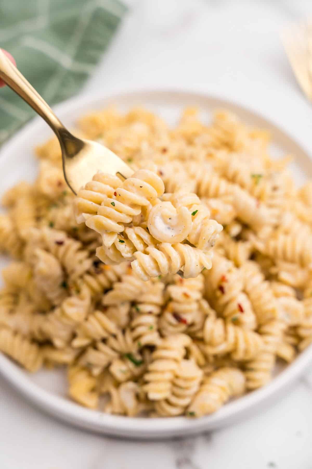 A fork lifting a bite of creamy rotini pasta from a plate, showing the cheesy sauce coating the noodles, with red pepper flakes and parsley as garnish.