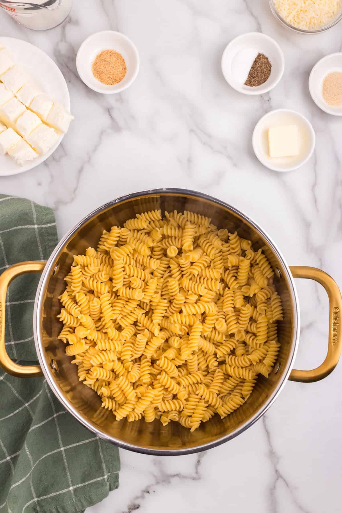 Cream cheese cubes, shredded cheese, and butter placed on top of uncooked rotini pasta inside a pot, ready for cooking.