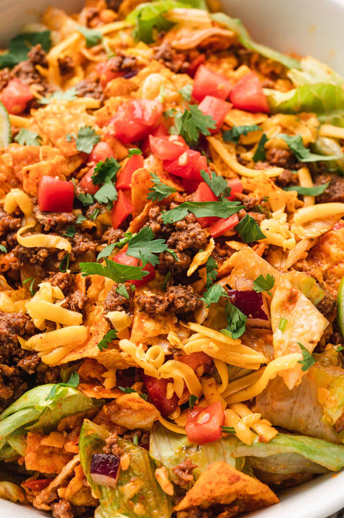 A close-up of Dorito taco salad, highlighting crispy crushed Doritos, seasoned ground beef, shredded cheddar cheese, chopped lettuce, diced tomatoes, and fresh cilantro. The vibrant colors and textures of the ingredients create a visually appealing dish with a mix of crunch, freshness, and savory flavors.