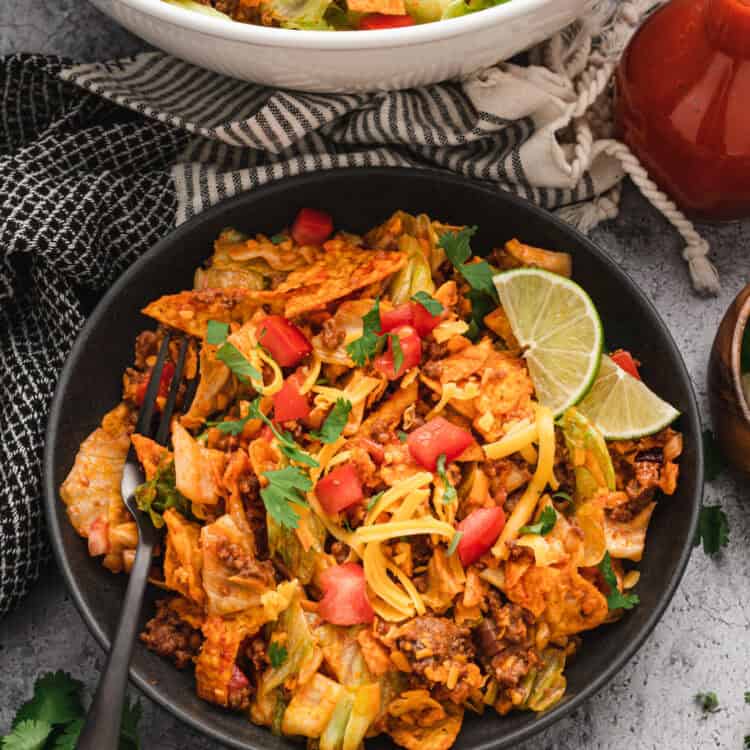 A plated serving of Dorito taco salad in a black bowl, garnished with fresh cilantro, diced tomatoes, shredded cheddar cheese, and lime wedges. The bowl sits on a textured gray surface, with a larger serving bowl in the background, a bottle of taco sauce, and fresh ingredients scattered around, creating a vibrant and appetizing presentation.